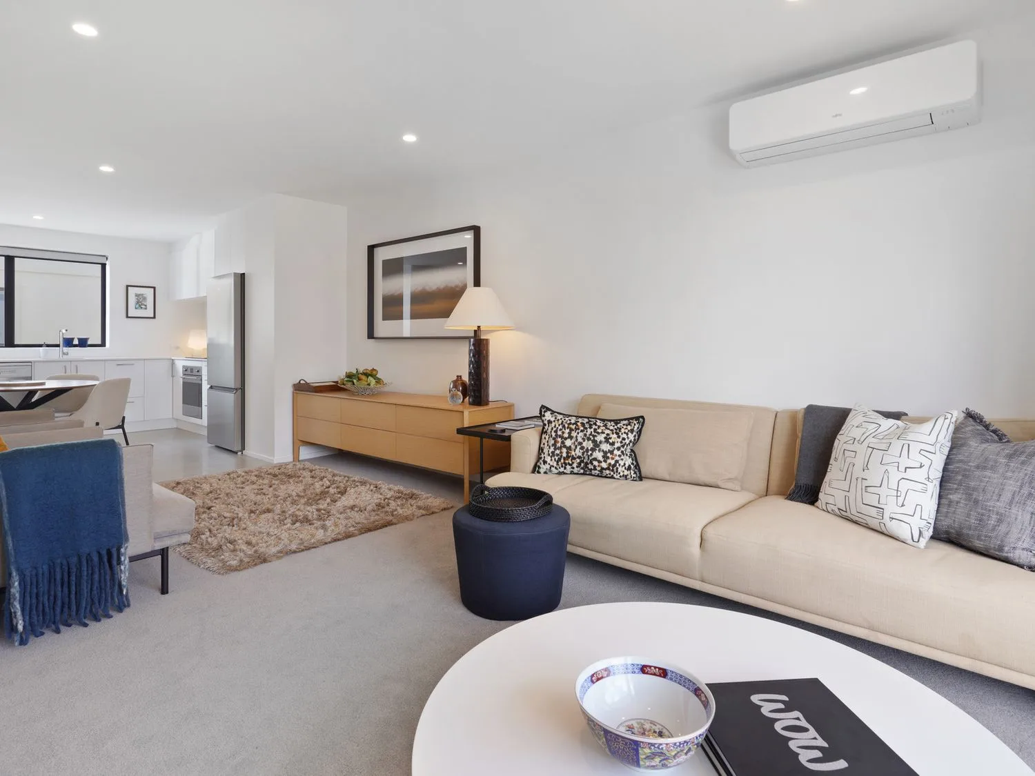 Una sala de estar con un sofá beige, cojines, una mesa de centro con un cuenco y un libro, un aparador de madera con una lámpara y una obra de arte, y una cocina al fondo en Modern Central Apartment - Christchurch Apartment, en Monavale