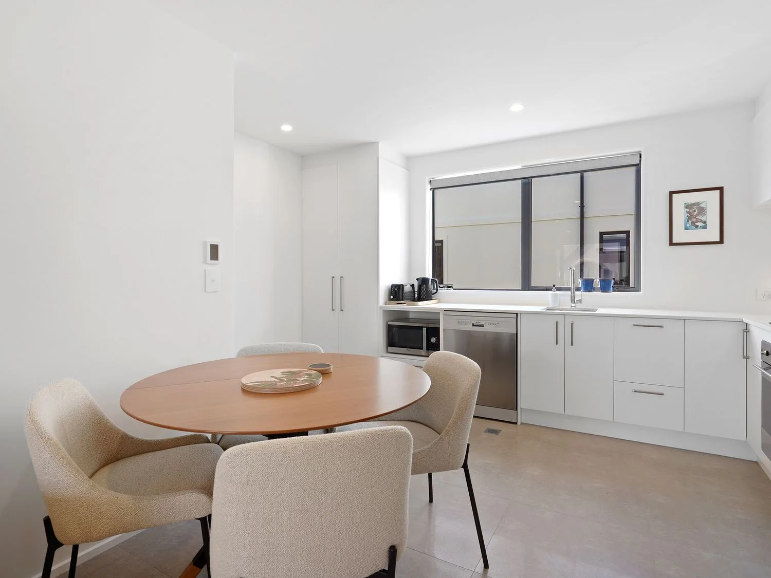 Una cocina luminosa tiene una mesa redonda con asientos junto a armarios blancos y una ventana amplia.