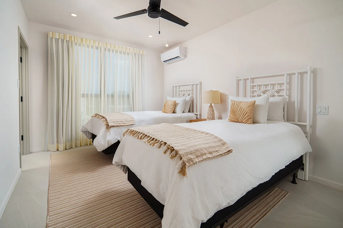 A bright bedroom with two beds, pale bedding, and floor-length curtains.