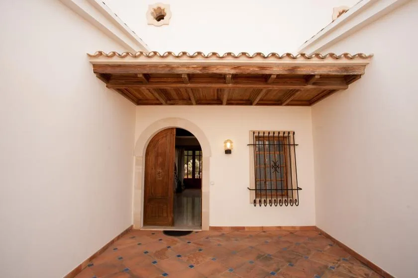 Un pequeño patio embaldosado tiene un toldo de madera, una puerta arqueada y una ventana con rejas.