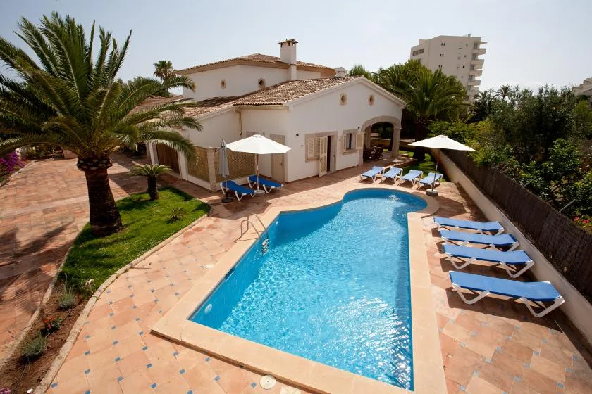 Una piscina junto a una casa blanca, con tumbonas en el patio.