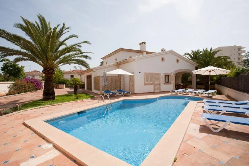 Una piscina junto a una casa blanca baja, con palmeras y tumbonas.