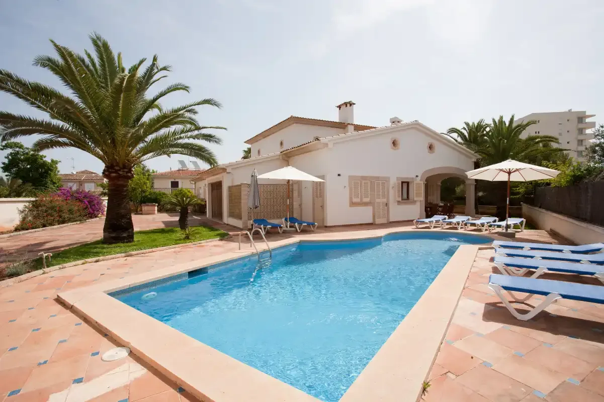 Una piscina está junto a una villa clara, con palmeras y tumbonas cerca.