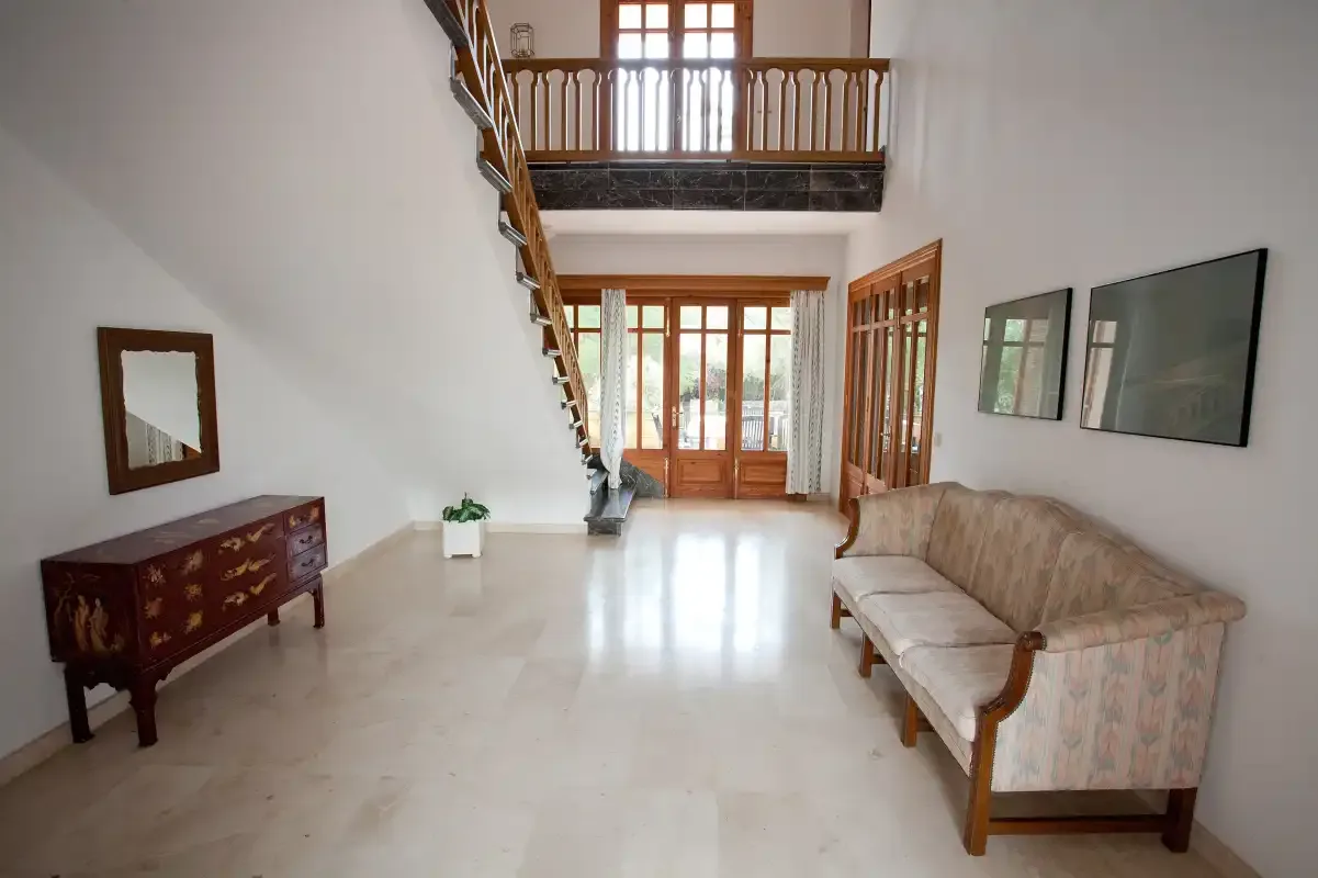 Una sala de estar luminosa con asientos, una escalera de madera y ventanas grandes.