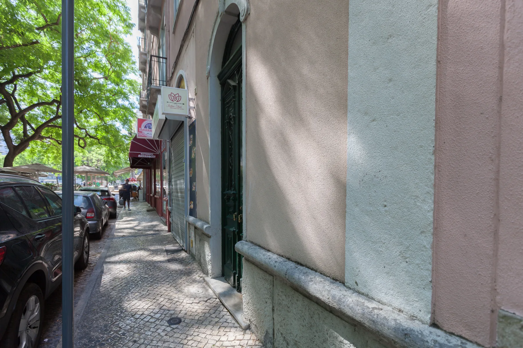 A tree-lined city sidewalk runs beside a row of parked cars and storefronts.