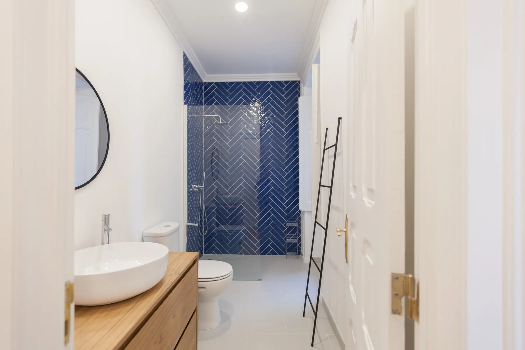 A narrow bathroom has a white sink, toilet, and dark blue tiled shower area.