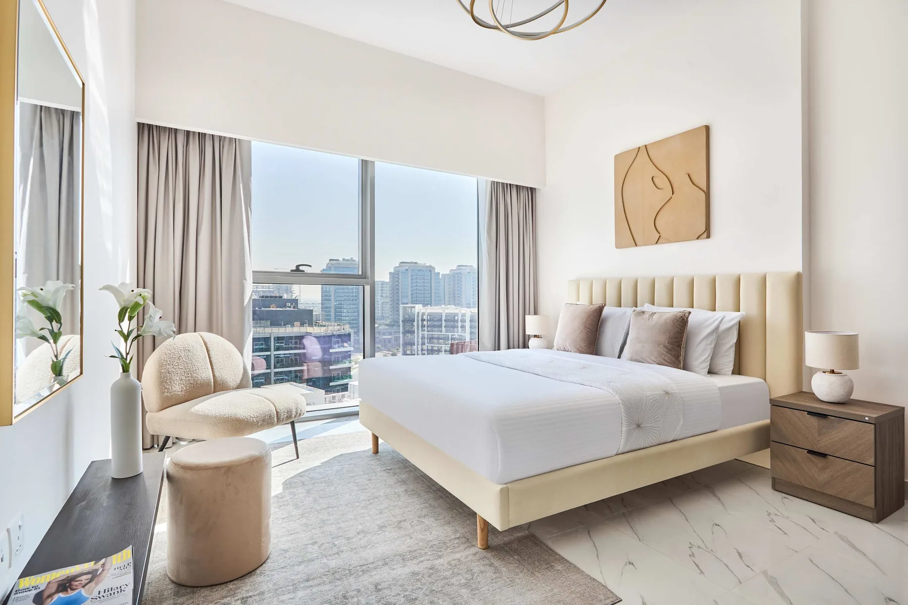 Un dormitorio luminoso con una cama tendida junto a un gran ventanal con vistas a edificios de la ciudad.
