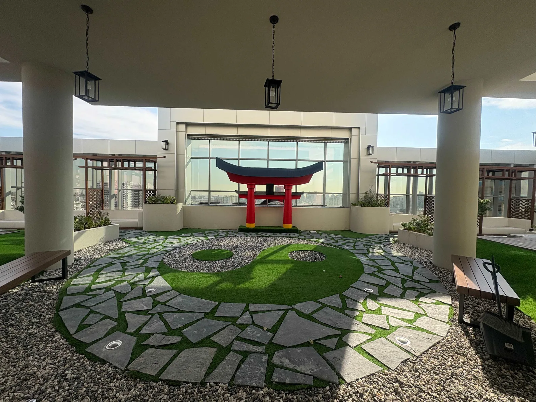 Un patio cubierto presenta una puerta ornamental roja y negra entre senderos de piedra y vegetación.
