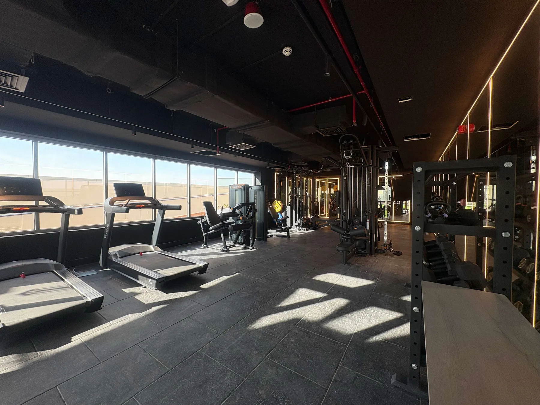 Un gimnasio con cintas de correr y equipo de entrenamiento de fuerza junto a grandes ventanas.