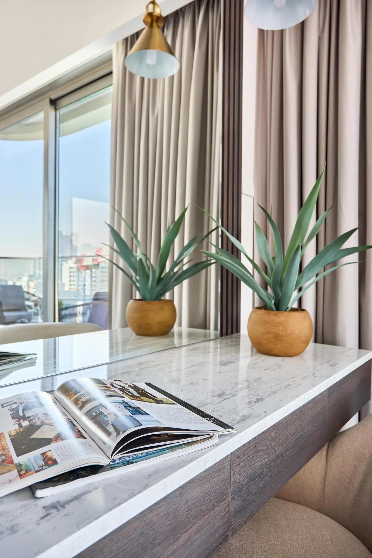 Un escritorio con superficie de mármol, una revista abierta y dos plantas junto a una ventana.