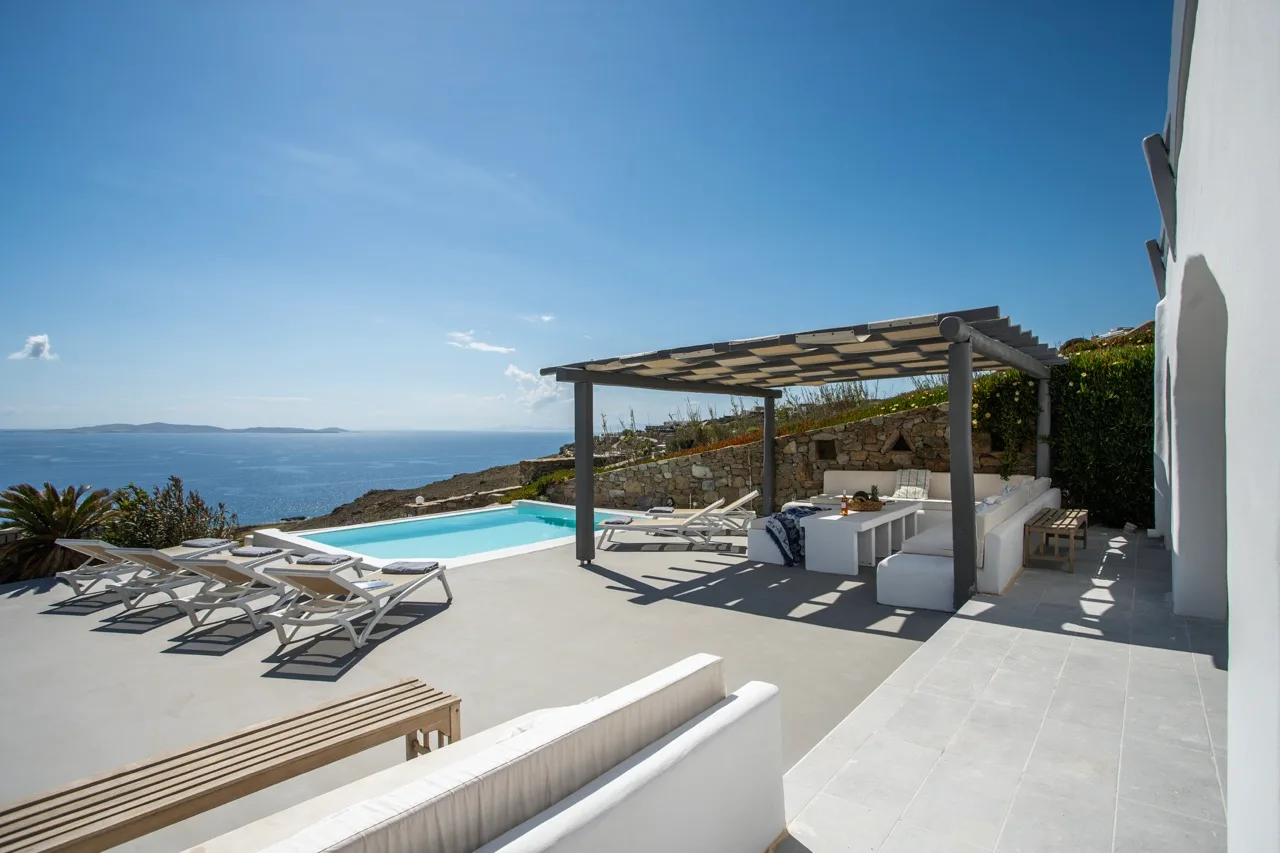 A sunny outdoor terrace overlooks the sea, with loungers beneath a pergola.