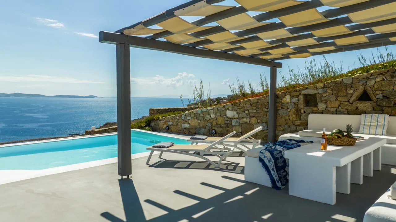 A poolside patio overlooks the sea, with loungers beneath a shaded pergola.