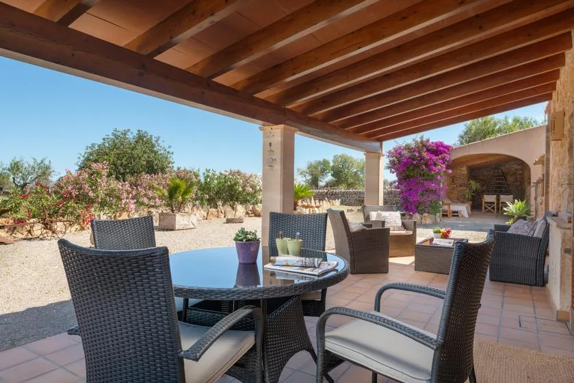 Un patio exterior cubierto tiene asientos oscuros alrededor de una mesa de vidrio y plantas con flores cerca.