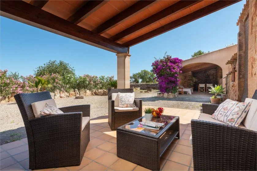 Un patio cubierto tiene asientos de mimbre alrededor de una mesa baja, con un jardín al fondo.
