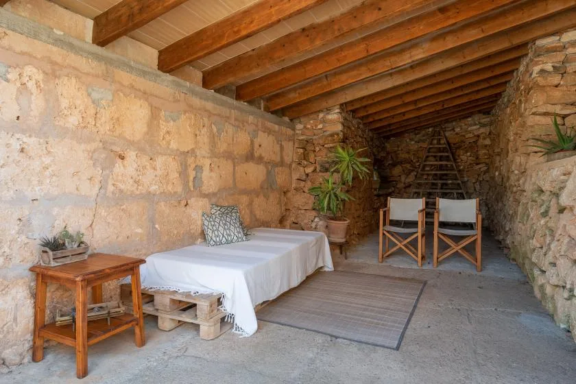 Una estancia rústica de piedra con una mesa, dos sillas y una cama cubierta con sábanas blancas.