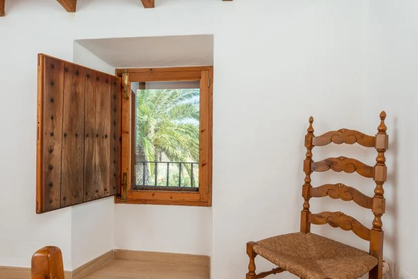 Una habitación luminosa con una ventana de madera que da a vegetación frondosa y una silla de madera.