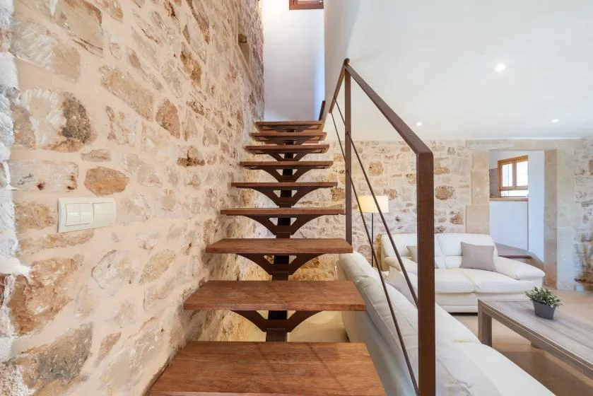 Una escalera de peldaños abiertos de madera sube junto a una pared de piedra en una sala luminosa.