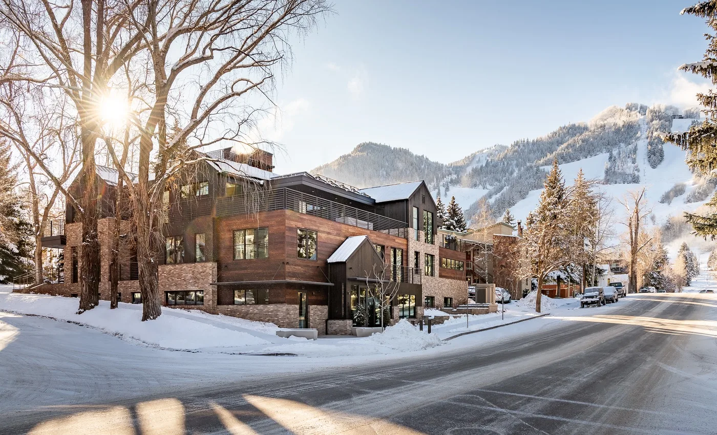 A modern lodge sits beside a snowy road, with wooded mountains in the background.