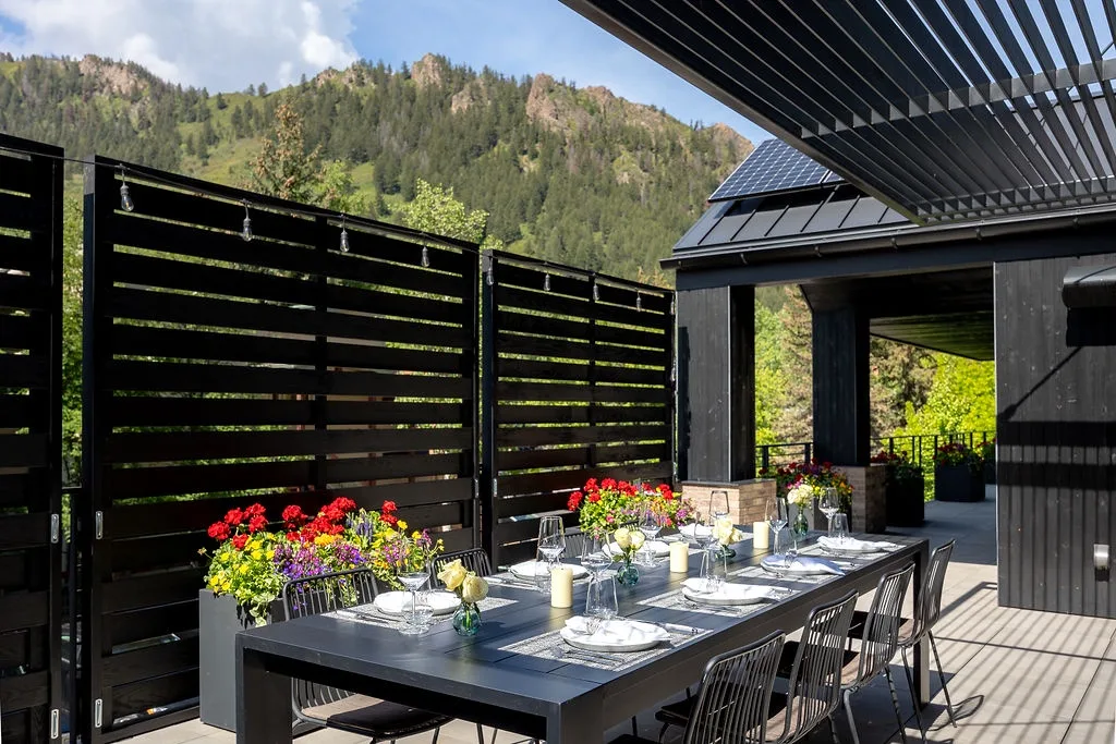 An outdoor dining table sits on a deck beside a black slatted screen, with wooded hills beyond.