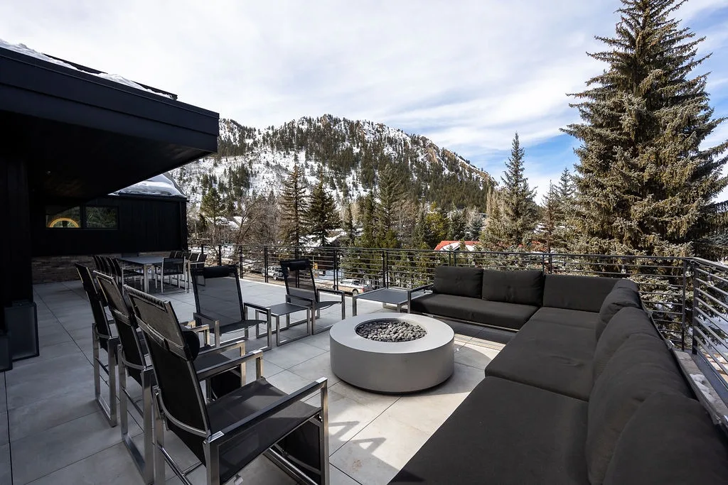 An outdoor terrace with seating and a round fire pit overlooks a wooded mountain.