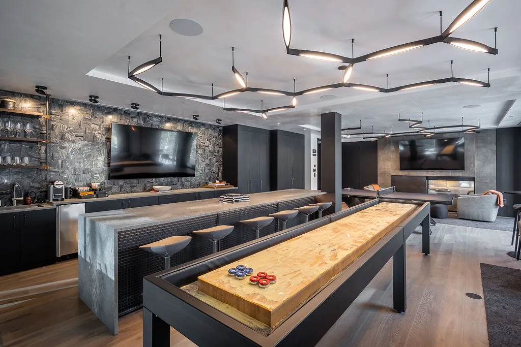 A modern game room features a long table and wall-mounted screens.