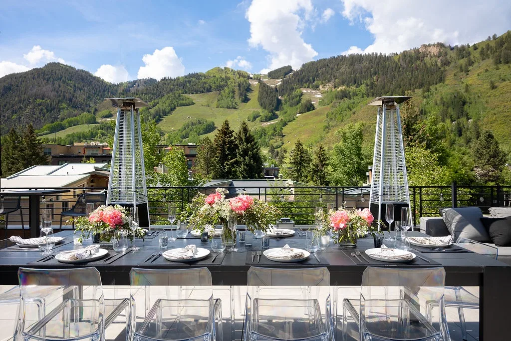 An outdoor dining table with clear seating overlooks green mountain slopes.