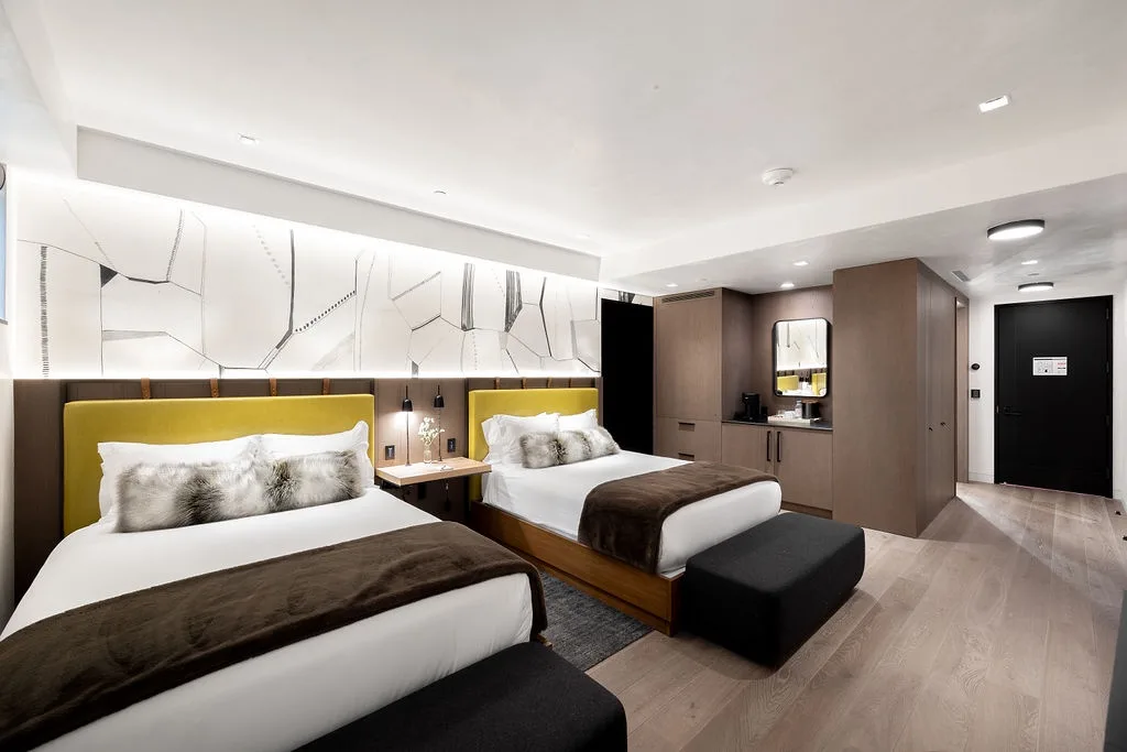 A modern bedroom with two beds, dark bedding, and wall-mounted lighting.
