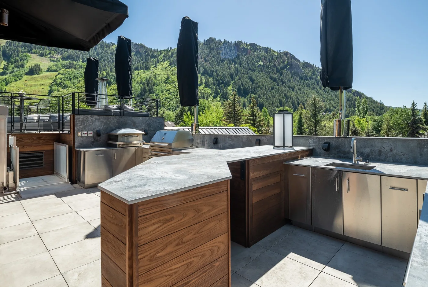An outdoor kitchen with wood cabinetry and a built-in grill overlooks a green mountainside.