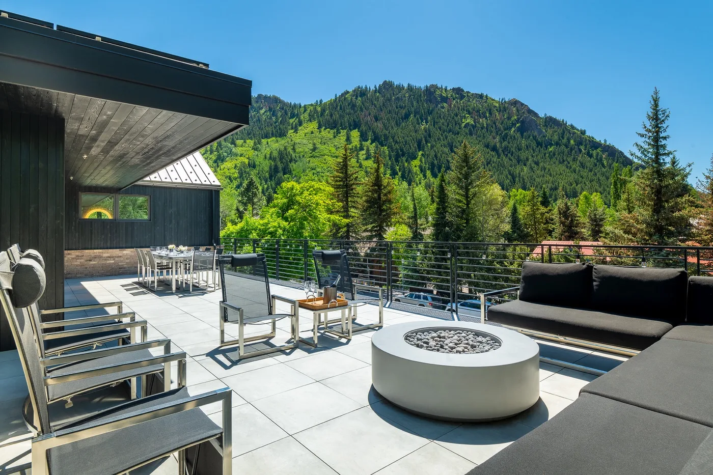 A rooftop terrace with outdoor seating and a round fire pit overlooks a forested mountain.