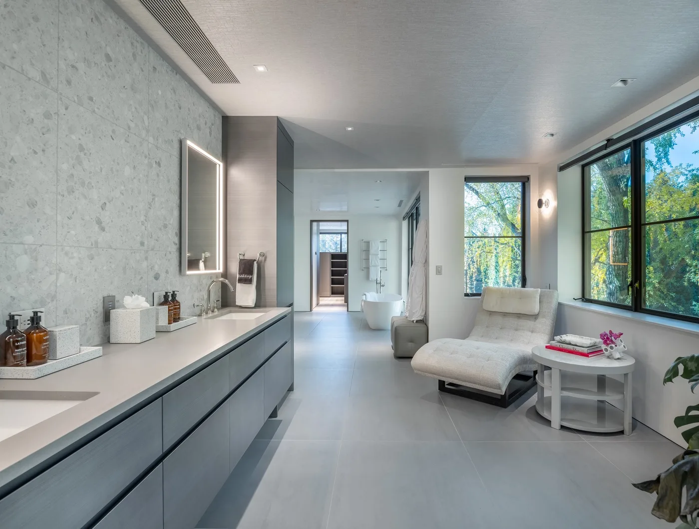 A bright bathroom with a long vanity, bathtub, and seating beside large windows.