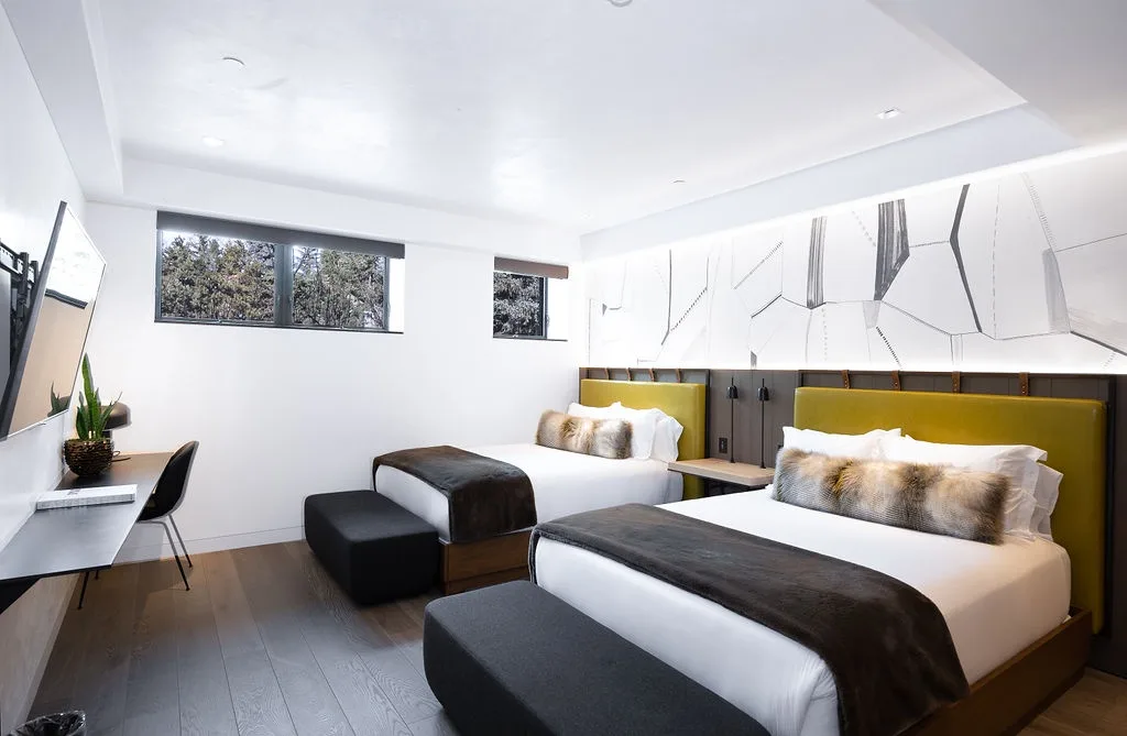 A bright bedroom with two beds, dark benches, and a desk by the window.