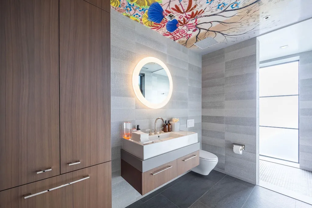 A modern bathroom with a vanity, illuminated round mirror, and colorful ceiling artwork.