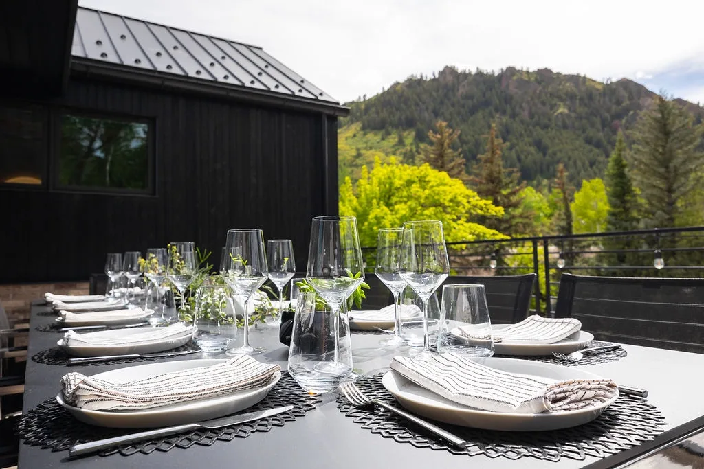 An outdoor dining table is set with plates and glassware beside a mountain view.