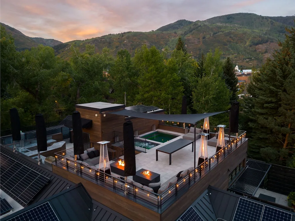 An elevated outdoor terrace with a pool and seating overlooks wooded hills at sunset.