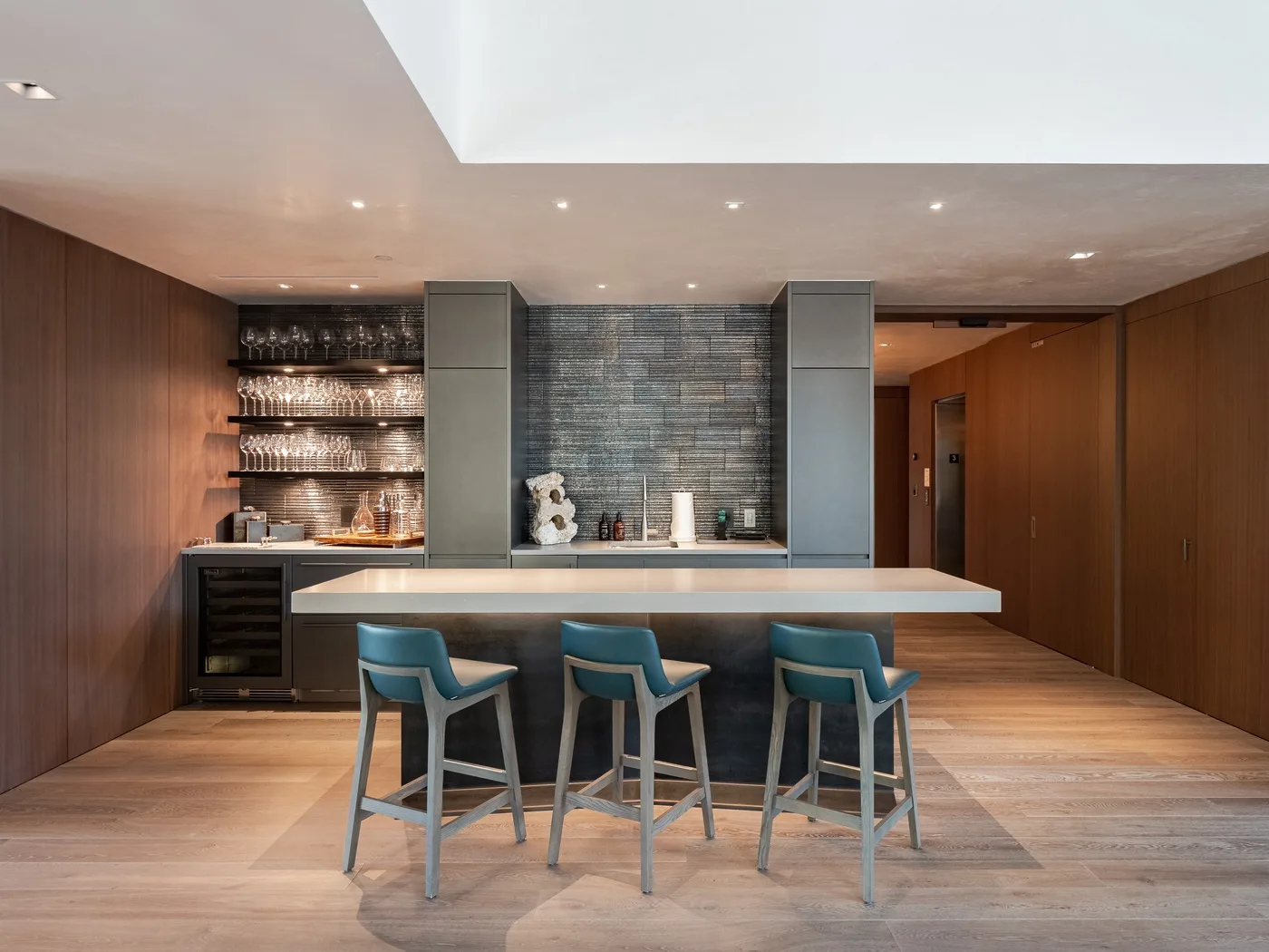 A modern bar counter with seating sits beside shelves of glassware.