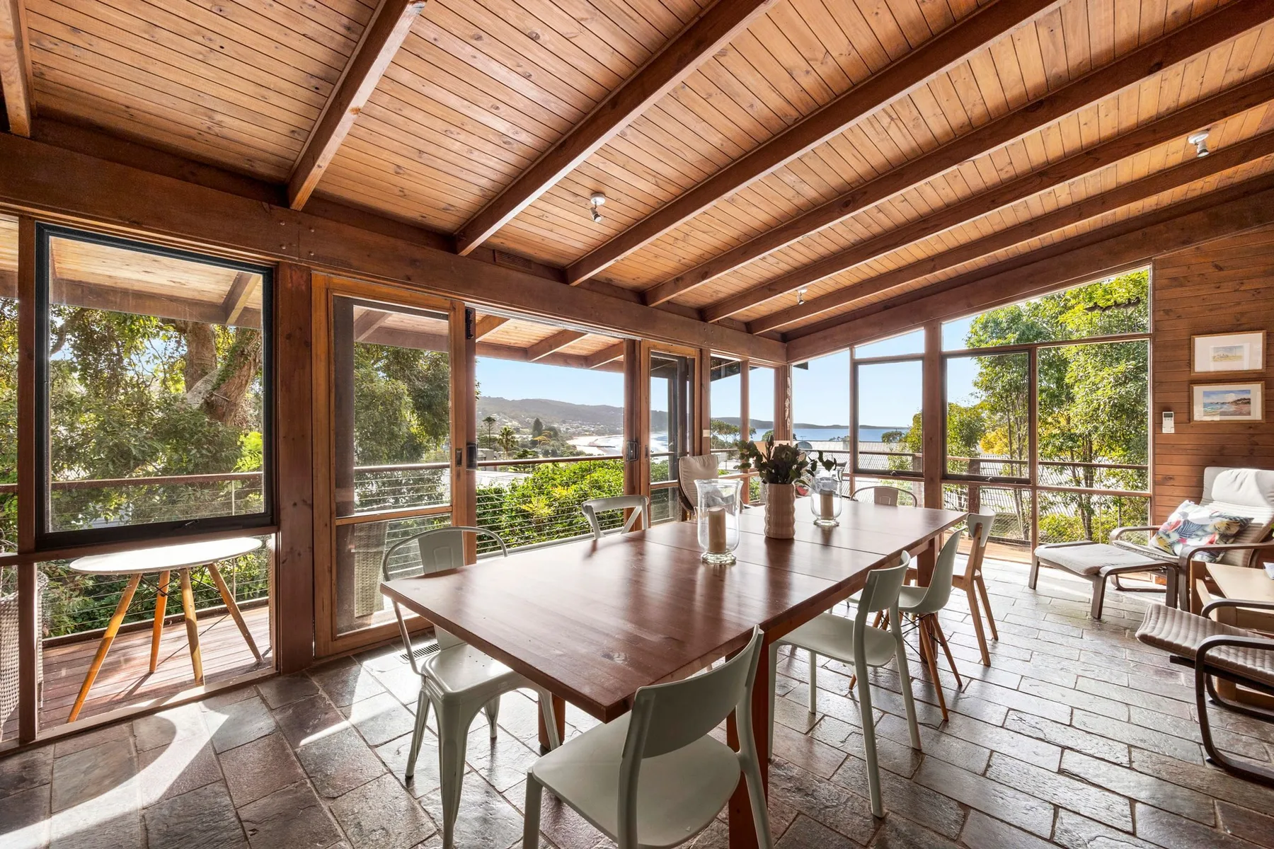 A bright dining area with a long table, seating, and wide windows overlooking the water.