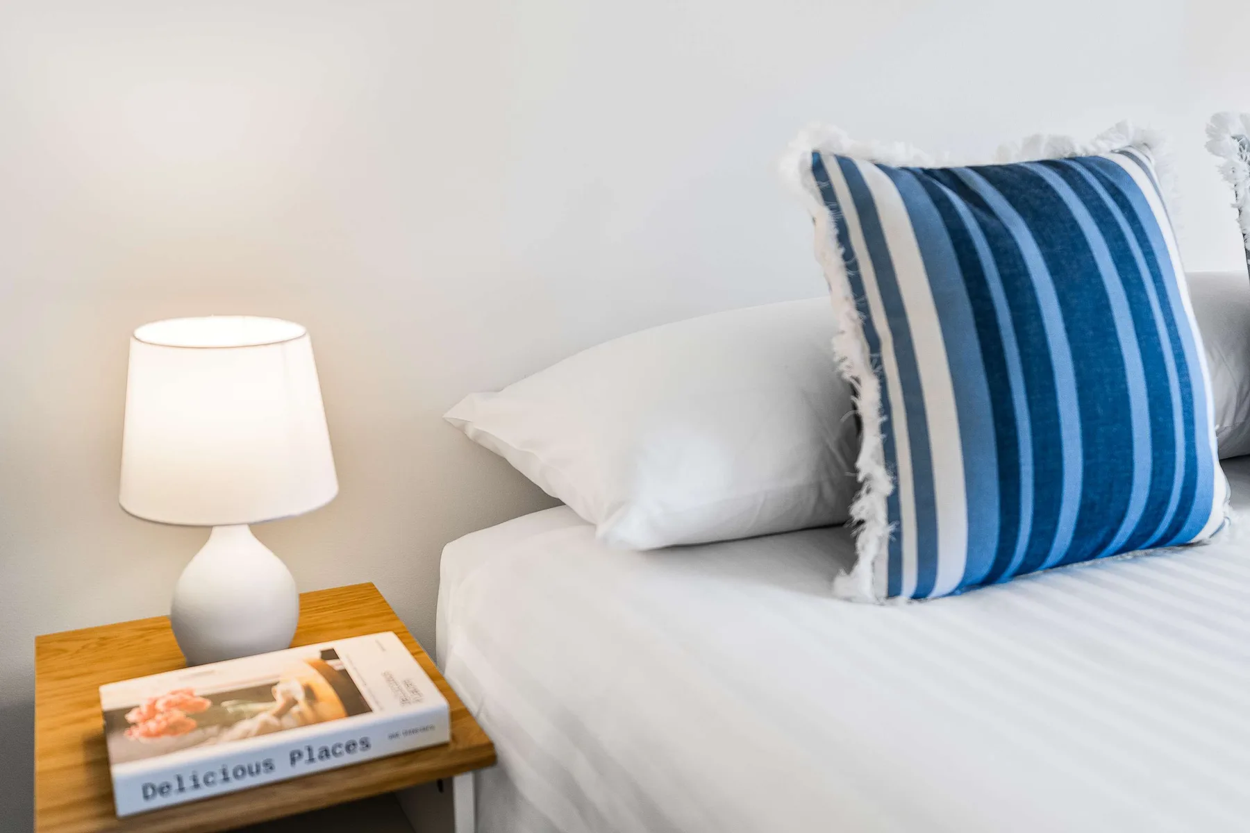 A bed with white bedding and a blue striped cushion beside a bedside table.