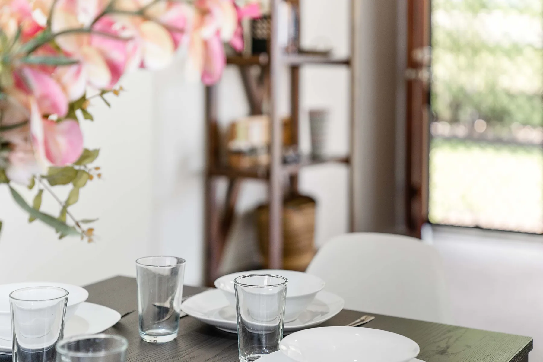 A dining table is set with plates and glasses beside a vase of flowers.