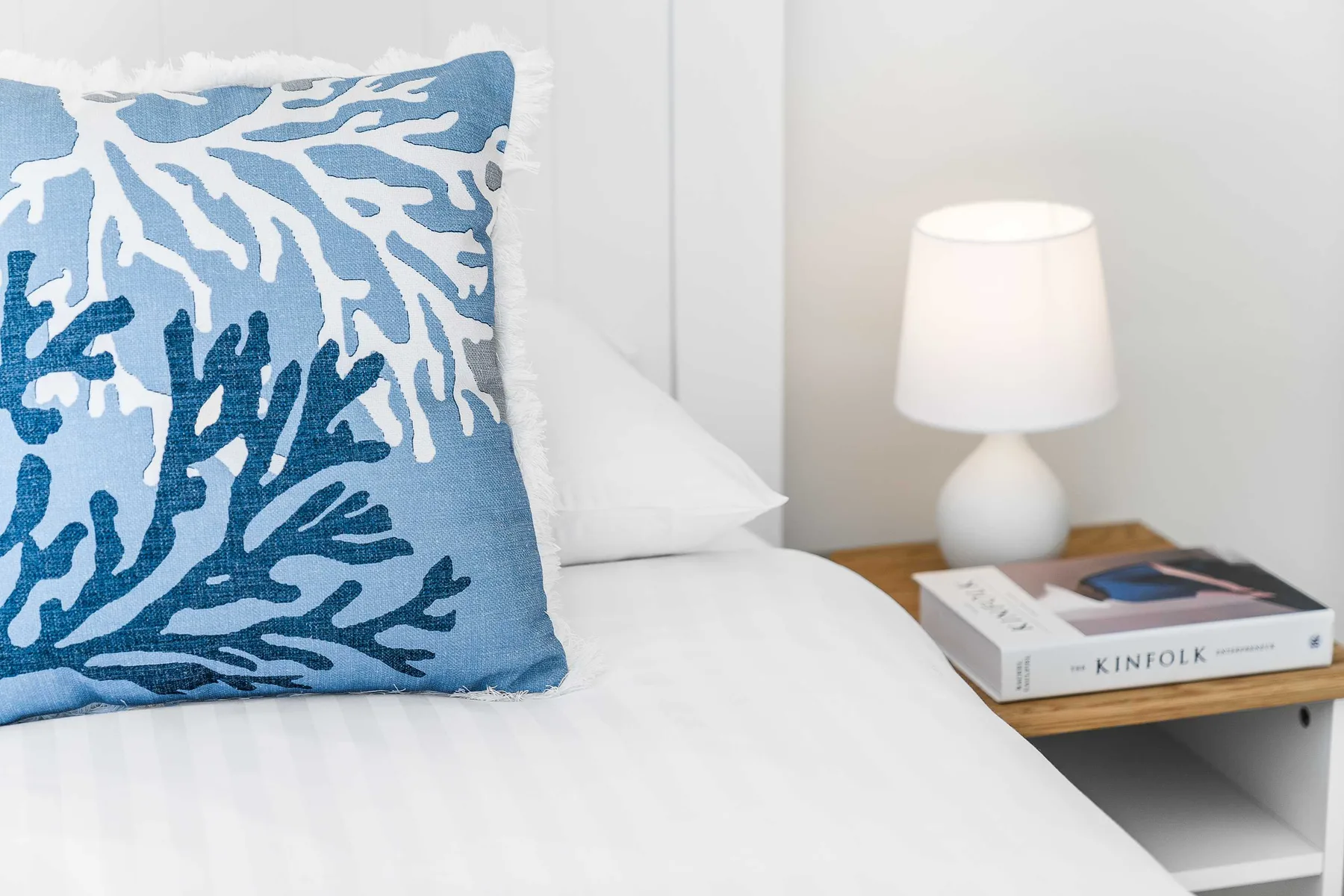 A bedroom bed with a blue patterned cushion beside a bedside table and lamp.