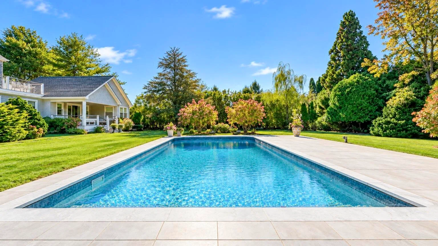 Una piscina exterior alargada está junto a un jardín paisajístico y una casa.