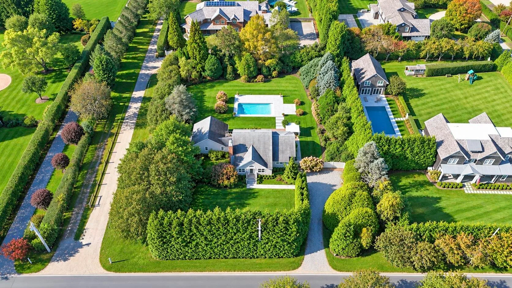 Vista aérea de casas, jardines y una piscina junto a una carretera arbolada.