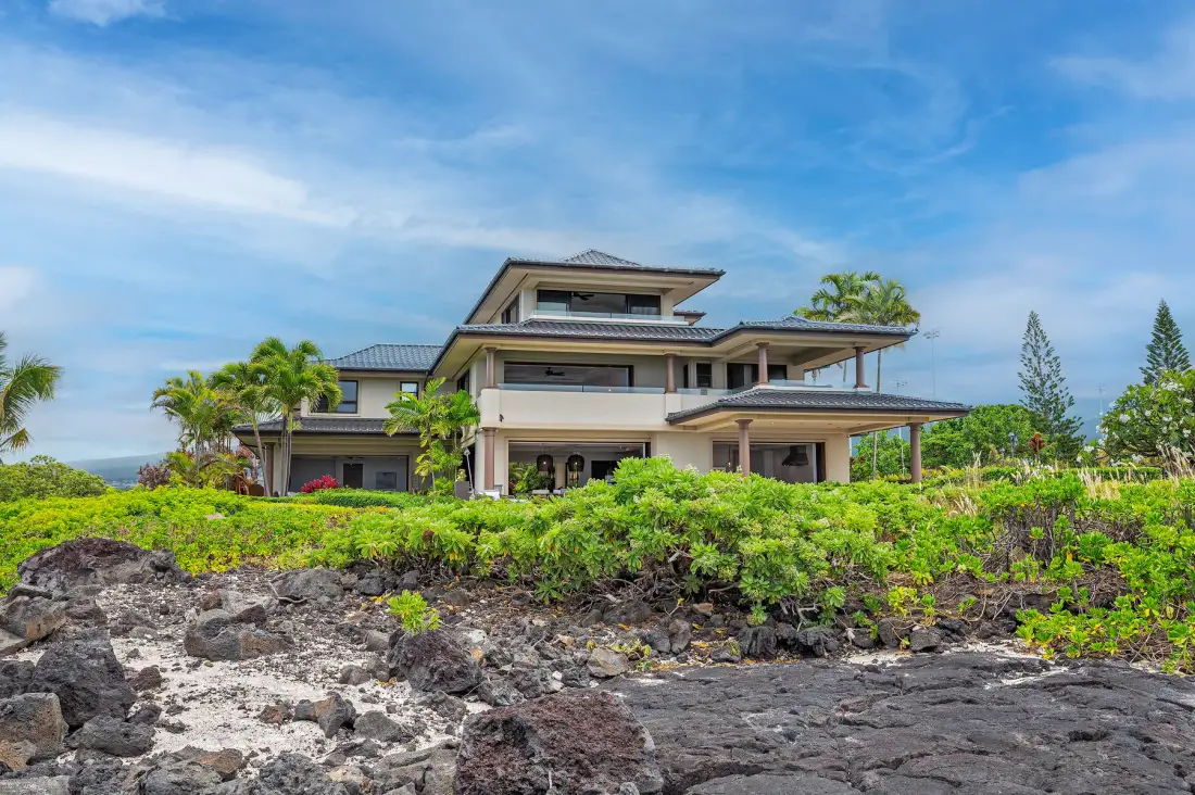 A large modern house stands behind low green shrubs on rocky ground.