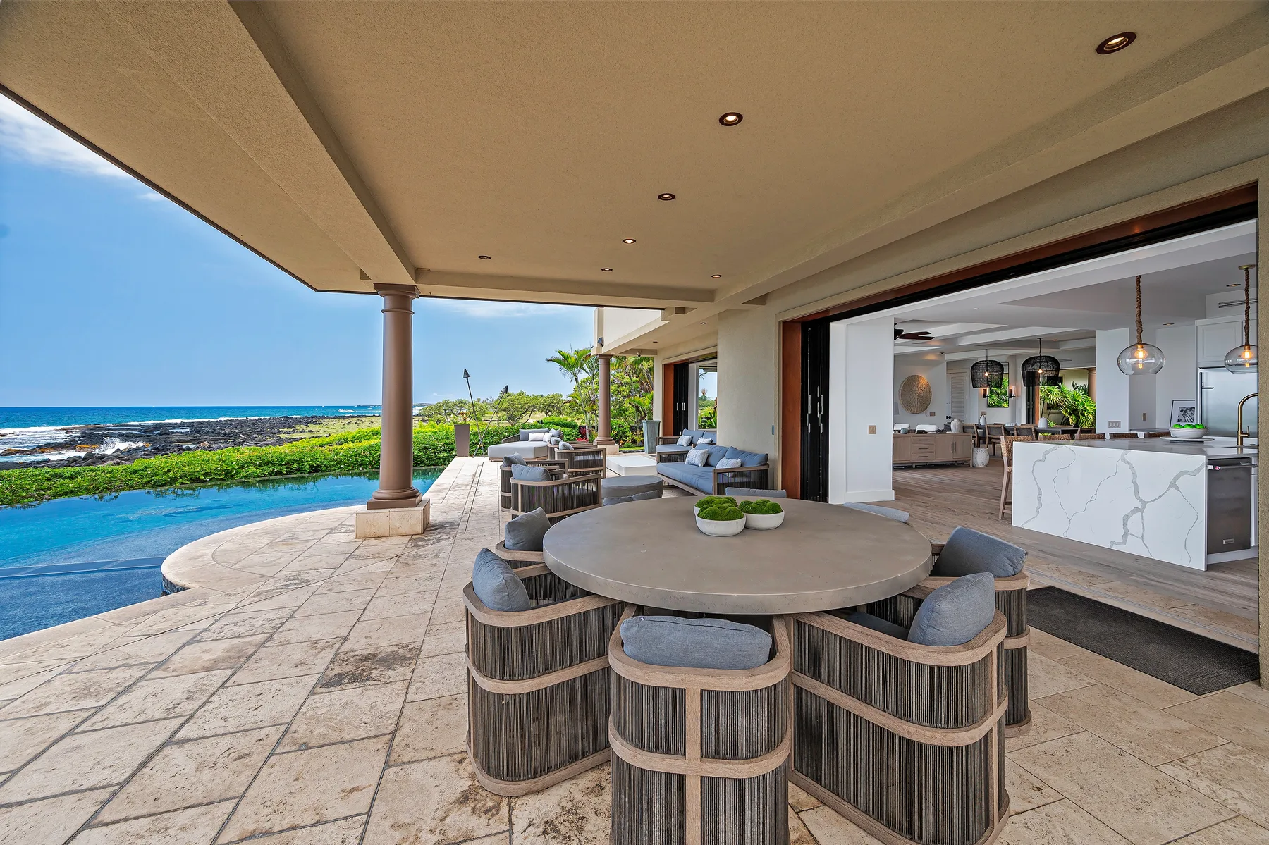 A covered outdoor dining area beside a pool, with an open kitchen beyond.