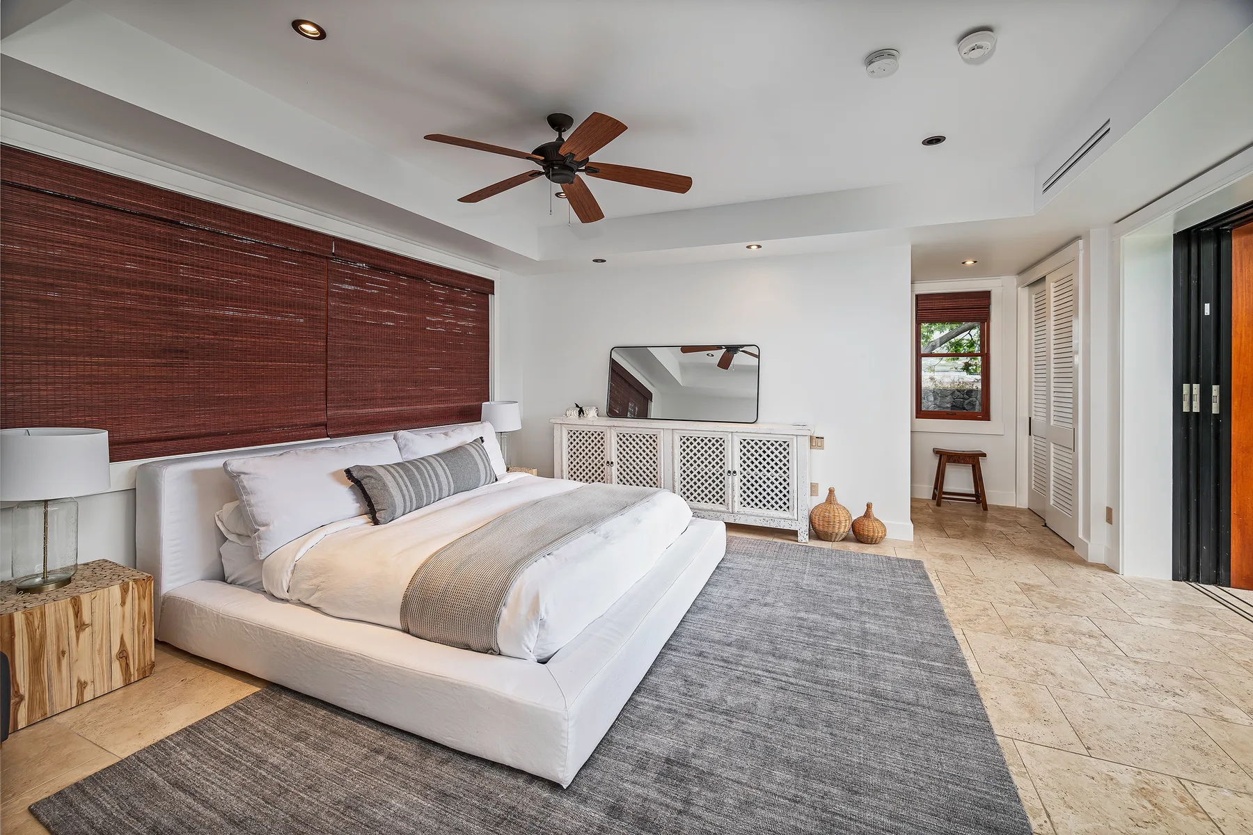 A bright bedroom with a low bed, gray rug, and wooden ceiling fan.