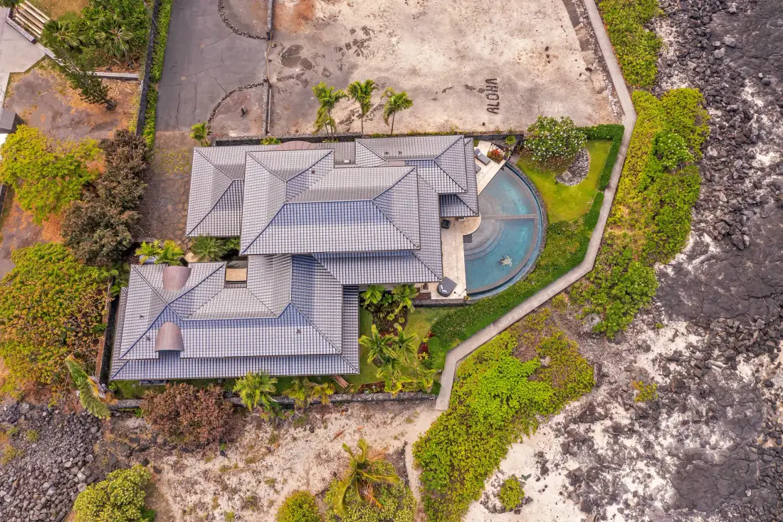 Aerial view of a tiled-roof building beside a curved swimming pool and rocky shoreline.
