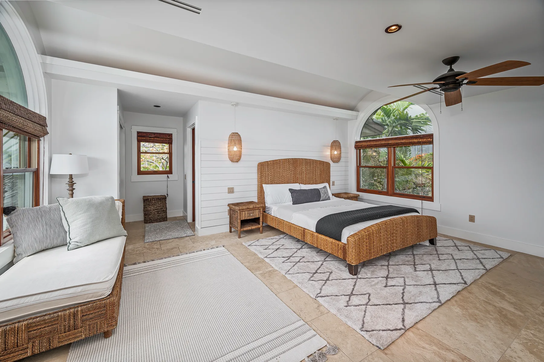 A bright bedroom with a bed, woven seating, and arched windows.