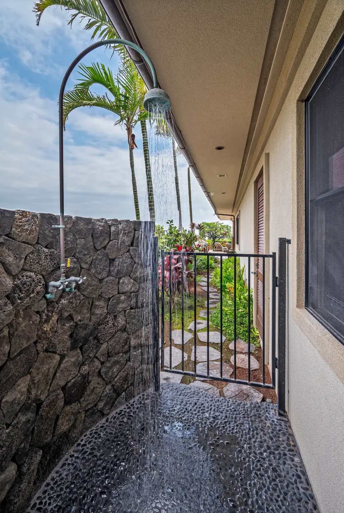 An outdoor shower area beside a stone wall, with tropical plants beyond a gate.