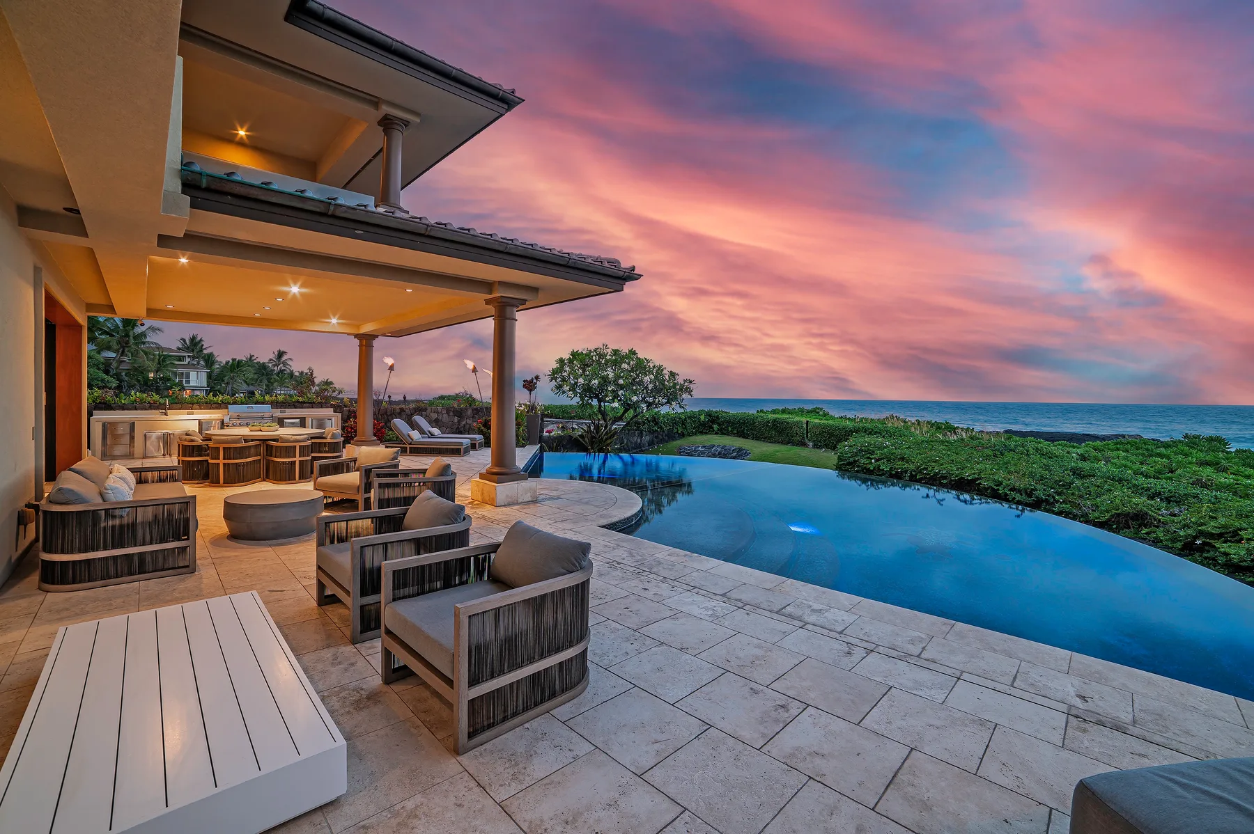 An outdoor patio with seating overlooks an infinity pool and the ocean at sunset.