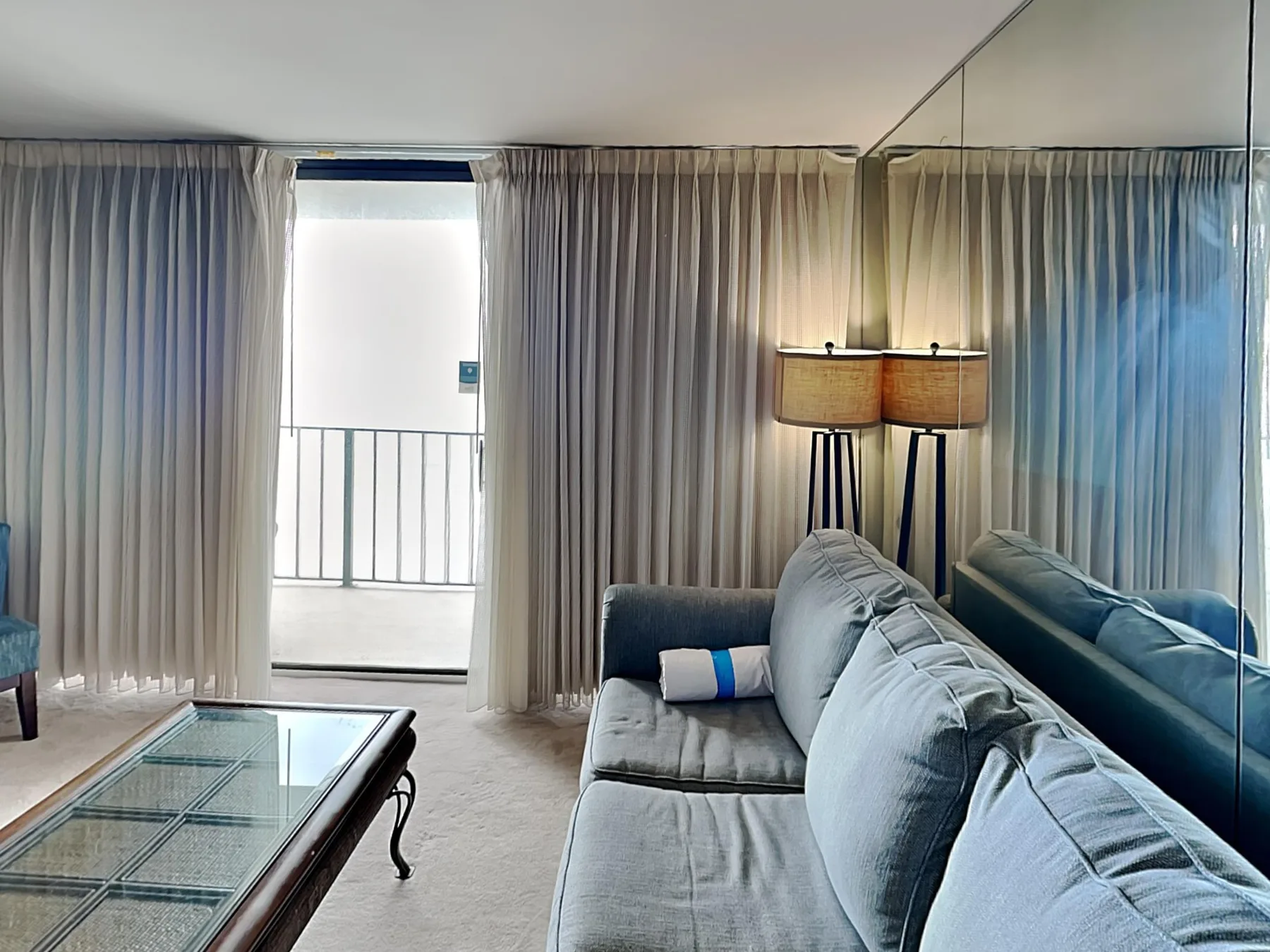 Una sala de estar con sofá gris, mesa de centro de vidrio y cortinas junto a una puerta de balcón.