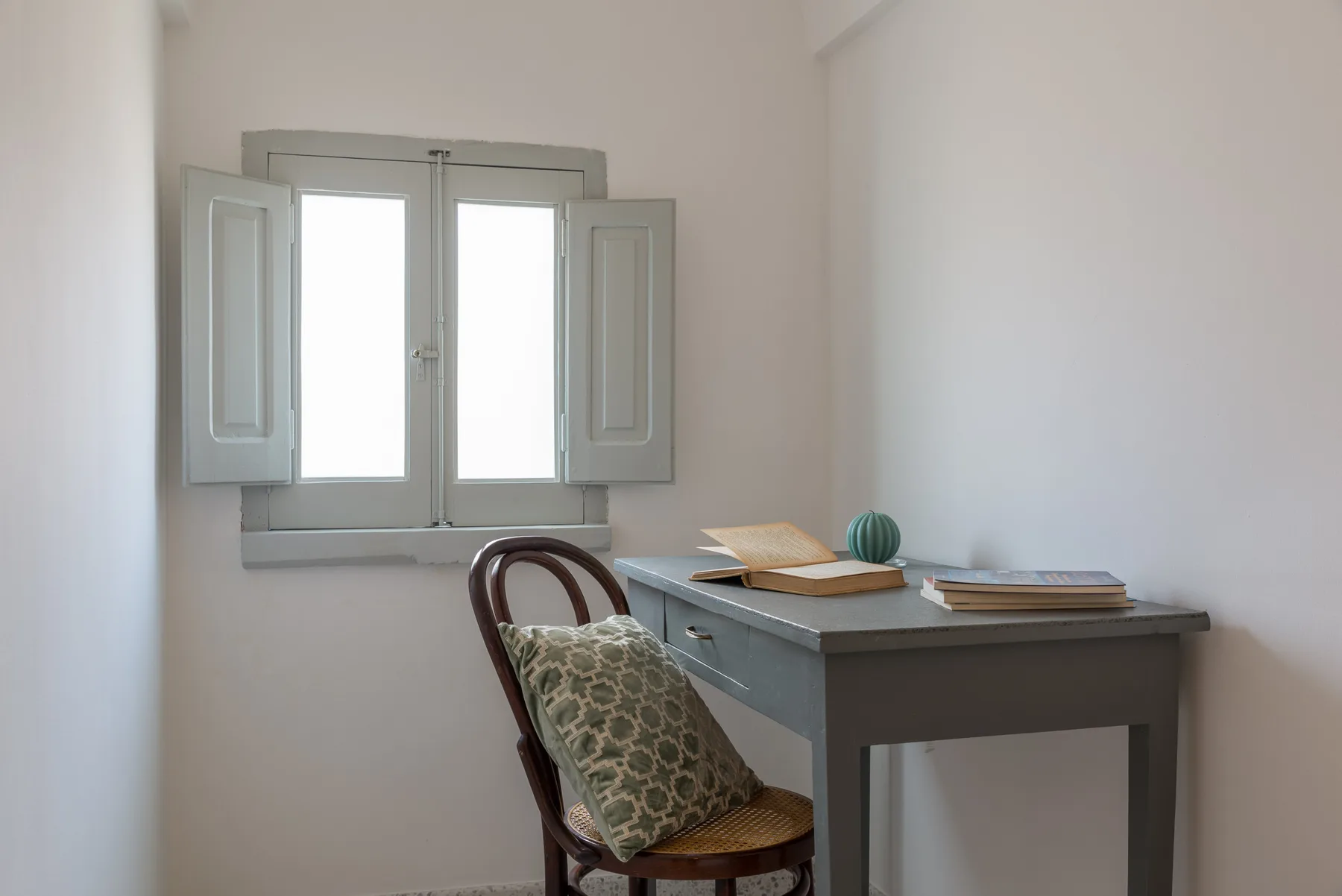 A small writing desk and chair sit beside a bright window in a simple room.